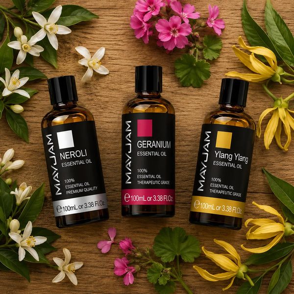 Három 100 ml-es illóolaj – neroli, geránium és ylang-ylang – rusztikus fa asztalon felülnézetből, virágokkal és zöld levelekkel körülvéve, prémium hangulatú természetes termékfotó.